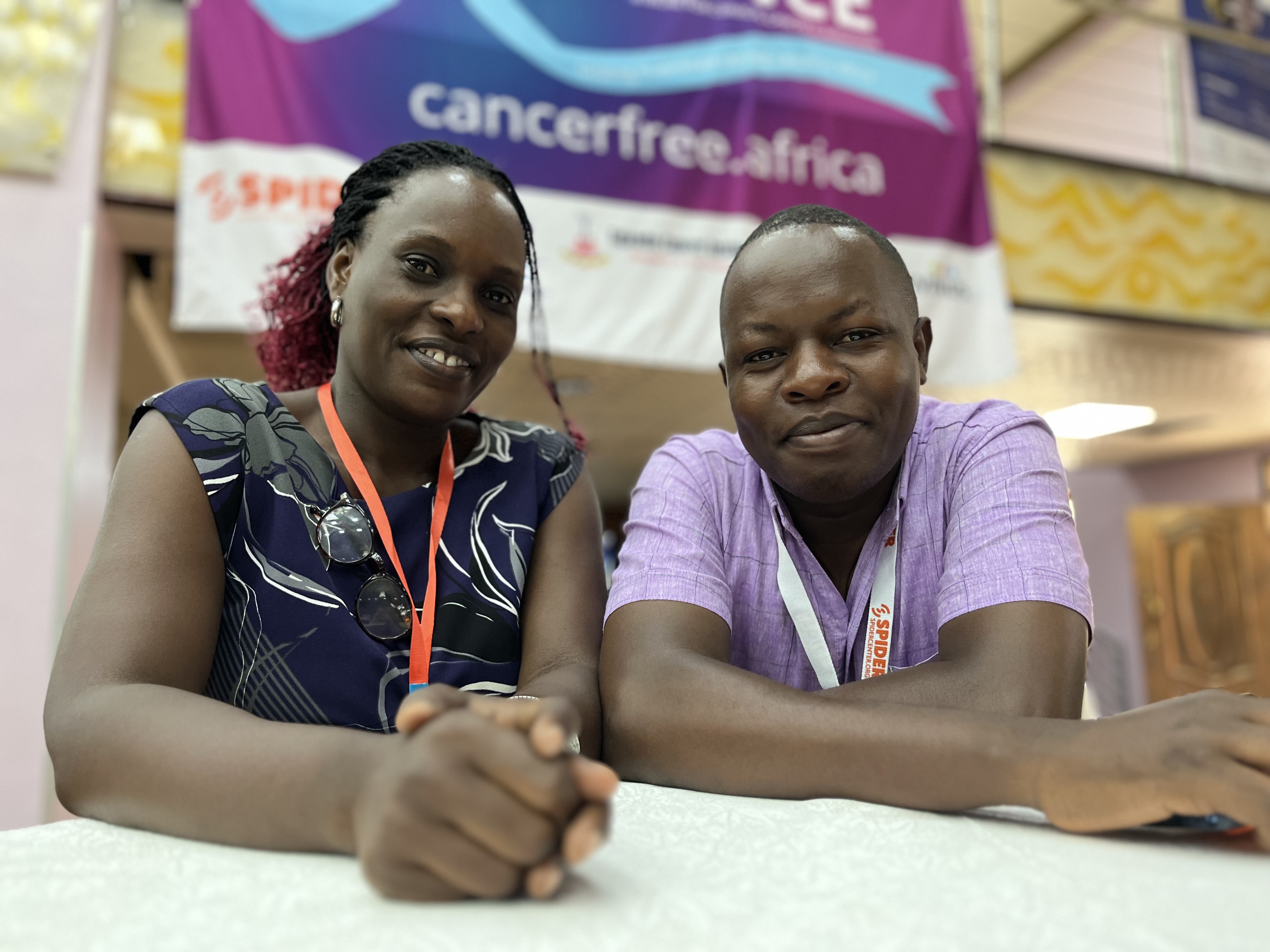 A smiling lady and a smiling gentlemen in professional clothing are standing by a table at a conference where a campaign banner is visible in the background with a weblink to cancerfree.africa.